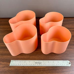 May include: Four peach-colored, cup-shaped containers with a wavy design. The containers are arranged on a wooden surface. A ruler is placed next to the containers for scale. The containers appear to be made of a 3D-printed material.