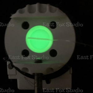 May include: A white plastic device with a green glowing light in the center. The device has a screw on the bottom and several small holes around the edge.