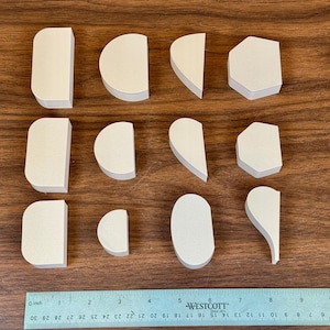 May include: An overhead shot shows twelve light-colored, geometric wooden shapes of varying sizes and forms. A ruler with inch measurements is at the bottom. The shapes are arranged on a brown wooden surface.