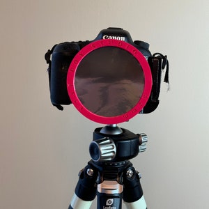 May include: A black Canon camera with a pink "FOX STUDIO" ring attached to the lens, mounted on a black tripod with a silver and blue Leofoto LS-324C tripod head.