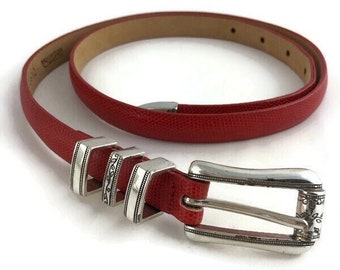 May include: A red belt with a textured surface and a silver buckle. The belt has a series of silver rings near the buckle. The belt is approximately 1 inch wide.
