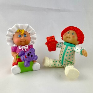 Könnte beinhalten: Zwei Cabbage Patch Kids Puppen. Eine Puppe trägt ein rosa Kleid und eine weiße Haube, hält einen lila Koala und sitzt in einem grünen und roten Spielzeug. Die andere Puppe trägt einen gepunkteten Strampler und hält einen roten Teddybären.