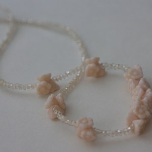 May include: A delicate necklace featuring a strand of small, clear beads. The necklace is adorned with several clusters of light pink, flower-shaped beads. The design is simple and elegant, suitable for various occasions.