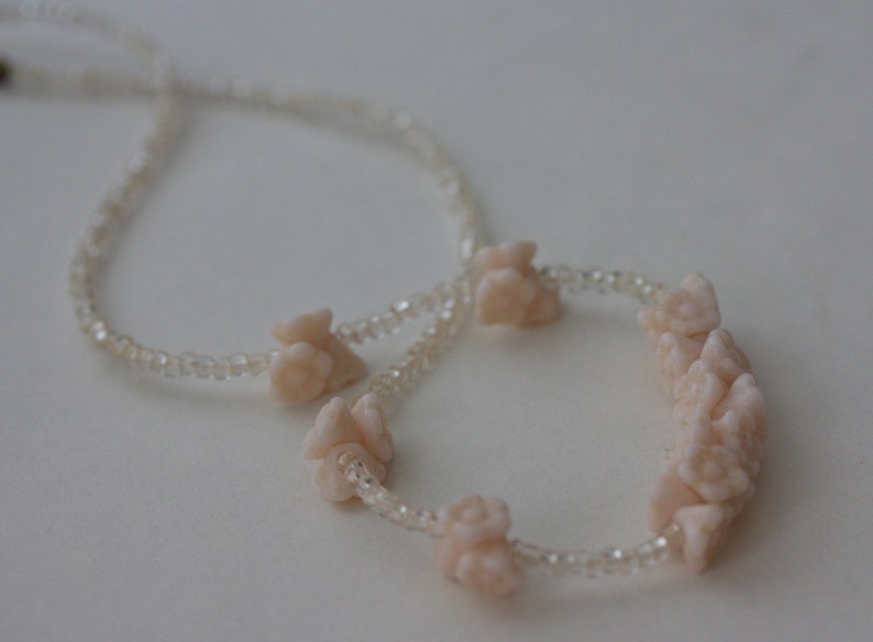 May include: A delicate necklace featuring a strand of small, clear beads. The necklace is adorned with several clusters of light pink, flower-shaped beads. The design is simple and elegant, suitable for various occasions.