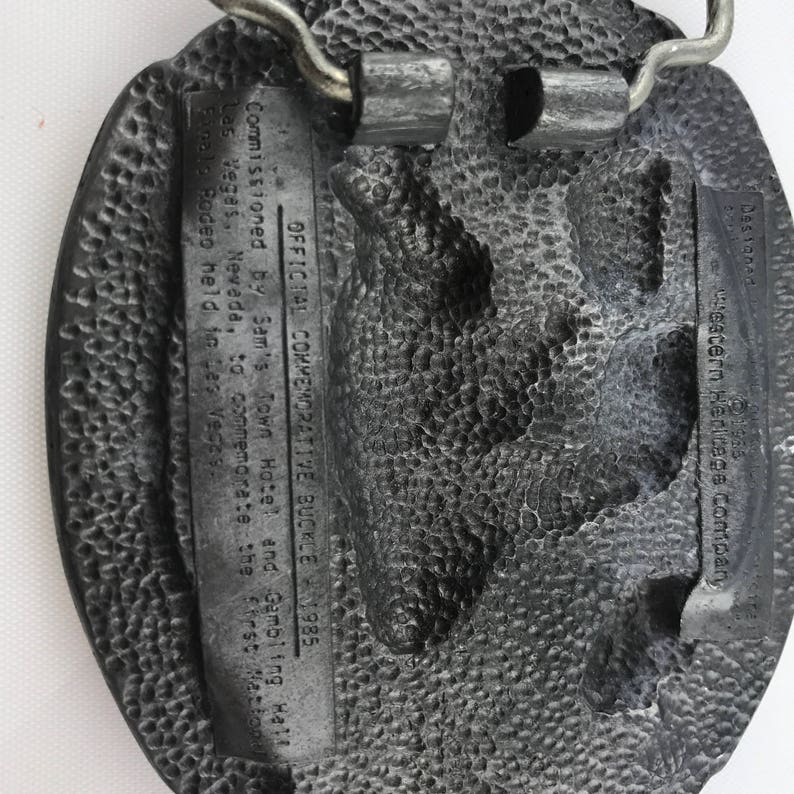 May include: A textured, oval-shaped metal belt buckle. The buckle has a detailed design and includes text that reads "OFFICIAL COMMEMORATIVE BUCKLE 1985" and "Las Vegas, Nevada". The buckle is a commemorative item.