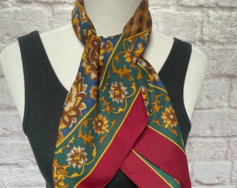 Può includere: Una sciarpa quadrata con un motivo floreale nei toni del verde, blu, marrone e oro, bordata di bordeaux. La sciarpa è esposta su un manichino, mettendo in risalto il suo design e i suoi colori.