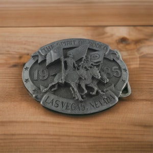 May include: Oval-shaped, pewter-colored belt buckle featuring a rodeo theme. The buckle displays two riders on horseback, holding flags, with the text "THE SPIRIT OF RODEO" and "LAS VEGAS, NEVADA". The year "1985" is also visible.