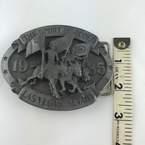 May include: A silver-colored belt buckle with the text "THE SPIRIT OF RODEO" and "LAS VEGAS, NEVADA". The buckle features a relief of two riders on horseback carrying flags, with the year "1985" displayed. The buckle measures approximately 3 inches.