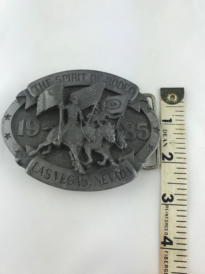 May include: A silver-colored belt buckle with the text "THE SPIRIT OF RODEO" and "LAS VEGAS, NEVADA". The buckle features a relief of two riders on horseback carrying flags, with the year "1985" displayed. The buckle measures approximately 3 inches.