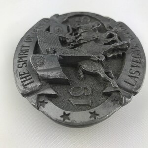 May include: A silver-colored belt buckle with a rodeo theme. The buckle features a cowboy riding a bucking bronco, an American flag, and the text "THE SPIRIT OF RODEO LAS VEGAS 19". The buckle has a circular shape with star accents.