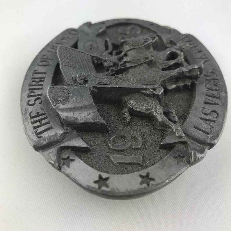 May include: A silver-colored belt buckle with a rodeo theme. The buckle features a cowboy riding a bucking bronco, an American flag, and the text "THE SPIRIT OF RODEO LAS VEGAS 19". The buckle has a circular shape with star accents.