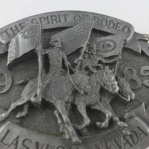 May include: A silver-colored belt buckle with the words "THE SPIRIT OF RODEO" and "LAS VEGAS NEVADA 85" engraved on it. The buckle features a detailed relief of three horses with riders carrying flags, including the American flag.