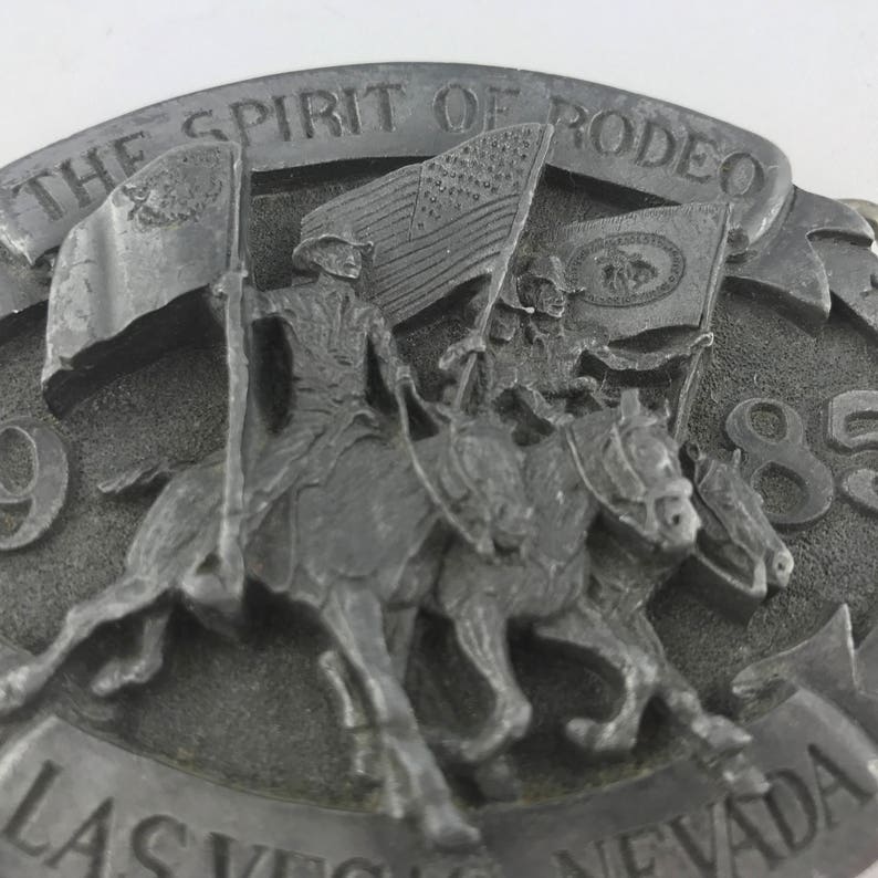 May include: A silver-colored belt buckle with the words "THE SPIRIT OF RODEO" and "LAS VEGAS NEVADA 85" engraved on it. The buckle features a detailed relief of three horses with riders carrying flags, including the American flag.