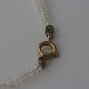 May include: Close-up of a delicate necklace with a gold-colored clasp. The necklace features small, clear beads strung together. The clasp is circular with a small lever for opening and closing. The background is a plain, light color.