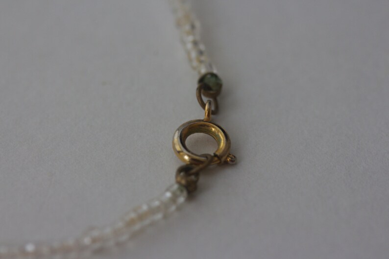 May include: Close-up of a delicate necklace with a gold-colored clasp. The necklace features small, clear beads strung together. The clasp is circular with a small lever for opening and closing. The background is a plain, light color.