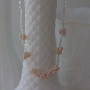 May include: A delicate necklace featuring small, peach-colored flower-shaped beads strung on a clear, beaded strand. The necklace is displayed against a backdrop of white, textured vases, creating a soft, feminine aesthetic.