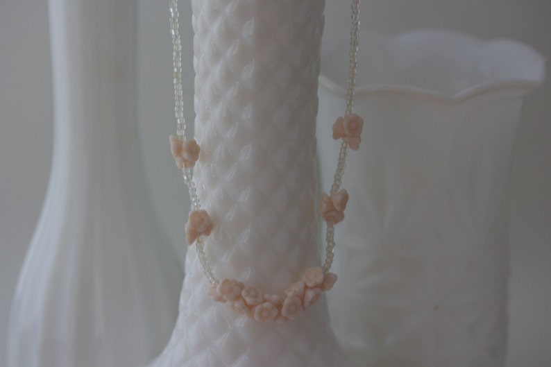 May include: A delicate necklace featuring small, peach-colored flower-shaped beads strung on a clear, beaded strand. The necklace is displayed against a backdrop of white, textured vases, creating a soft, feminine aesthetic.