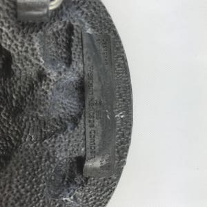 May include: A textured, oval-shaped, silver-colored belt buckle. The buckle has a rectangular inset with the text "Designed by Western Heritage Company" and other text. The buckle has a rugged, worn appearance.