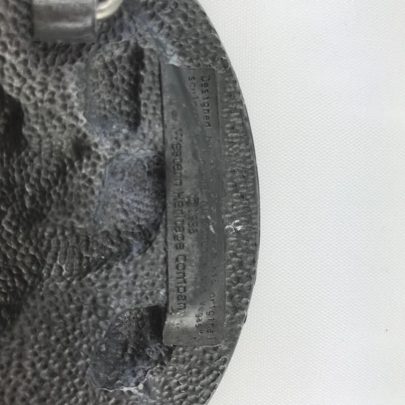 May include: A textured, oval-shaped, silver-colored belt buckle. The buckle has a rectangular inset with the text "Designed by Western Heritage Company" and other text. The buckle has a rugged, worn appearance.