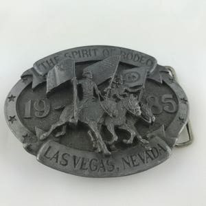 May include: A silver-colored belt buckle with a rodeo theme. The buckle features a relief of riders on horseback carrying flags, with the text "THE SPIRIT OF RODEO", "1985", and "LAS VEGAS, NEVADA". The buckle has an oval shape with star accents.