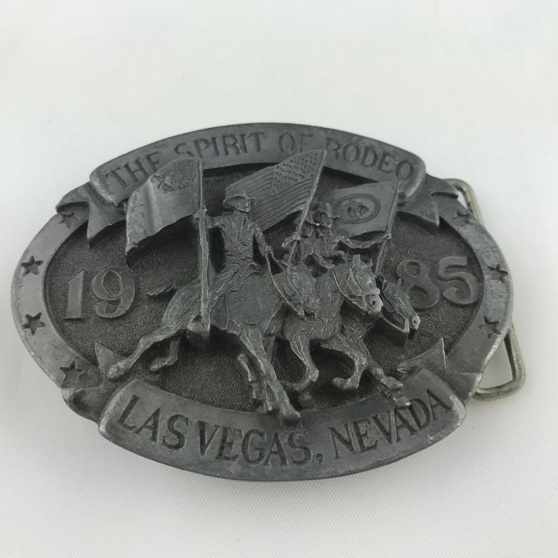 May include: A silver-colored belt buckle with a rodeo theme. The buckle features a relief of riders on horseback carrying flags, with the text "THE SPIRIT OF RODEO", "1985", and "LAS VEGAS, NEVADA". The buckle has an oval shape with star accents.