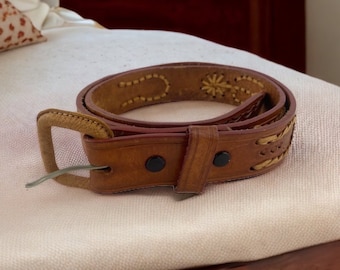 May include: A brown leather belt with decorative stitching and a rectangular buckle. The belt features a series of stitched patterns and a classic design. The belt is a medium brown color and has two black snaps.