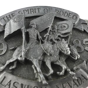 May include: A silver-toned belt buckle with the text "THE SPIRIT OF RODEO" and "LAS VEGAS NEVADA 85". The buckle features a detailed relief of riders on horseback carrying flags, including the American flag.