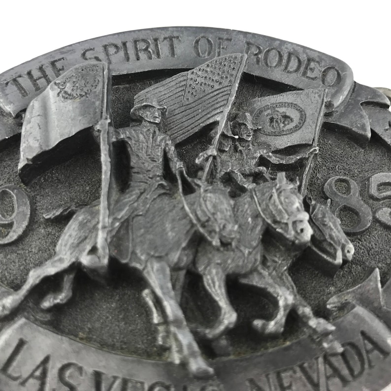 May include: A silver-toned belt buckle with the text "THE SPIRIT OF RODEO" and "LAS VEGAS NEVADA 85". The buckle features a detailed relief of riders on horseback carrying flags, including the American flag.
