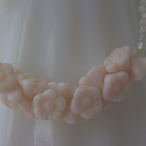 May include: A delicate necklace featuring a strand of pale pink flower-shaped beads. The flowers are clustered together, interspersed with clear, small beads. The necklace is set against a soft, white background.