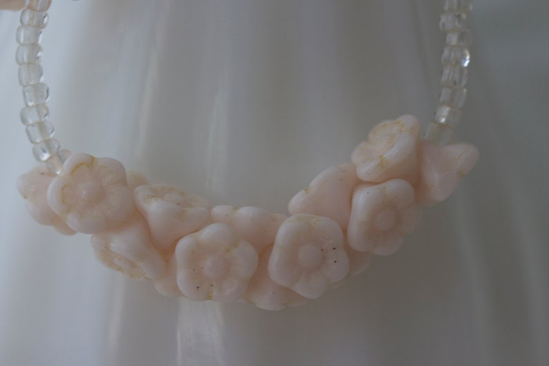 May include: A delicate necklace featuring a strand of pale pink flower-shaped beads. The flowers are clustered together, interspersed with clear, small beads. The necklace is set against a soft, white background.