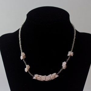 May include: A delicate necklace featuring small, light pink flower-shaped beads. The flowers are interspersed with clear, sparkling beads, creating a subtle contrast. The necklace is displayed on a black mannequin, highlighting its design.