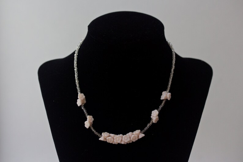 May include: A delicate necklace featuring small, light pink flower-shaped beads. The flowers are interspersed with clear, sparkling beads, creating a subtle contrast. The necklace is displayed on a black mannequin, highlighting its design.
