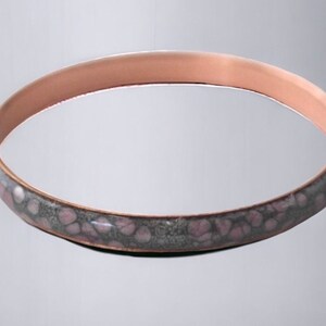 May include: A copper-coloured bangle bracelet. The decorative band features a pattern of pink and grey spots. The bracelet has a smooth, polished finish and a simple, elegant design. Jewellery.