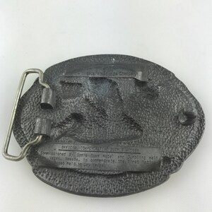 May include: A vintage, oval-shaped belt buckle with a textured, dark gray finish. The buckle features a silver-colored metal bar and a detailed relief design. Text on the buckle reads "OFFICIAL COMMEMORATIVE BUCKLE 1985" and "Sam's Town Hotel and Gambling Hall, Las Vegas, Nevada."