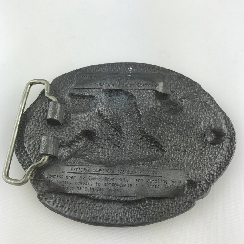 May include: A vintage, oval-shaped belt buckle with a textured, dark gray finish. The buckle features a silver-colored metal bar and a detailed relief design. Text on the buckle reads "OFFICIAL COMMEMORATIVE BUCKLE 1985" and "Sam's Town Hotel and Gambling Hall, Las Vegas, Nevada."