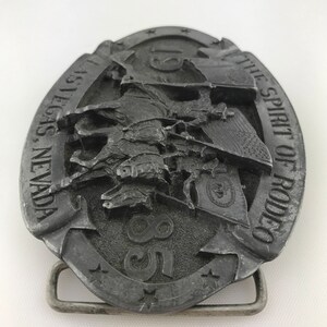 May include: A silver-colored metal belt buckle with a rodeo theme. The oval buckle features a detailed relief of a cowboy riding a bucking bronco, with the text "LAS VEGAS, NEVADA" and "THE SPIRIT OF RODEO". The number "85" is also visible.