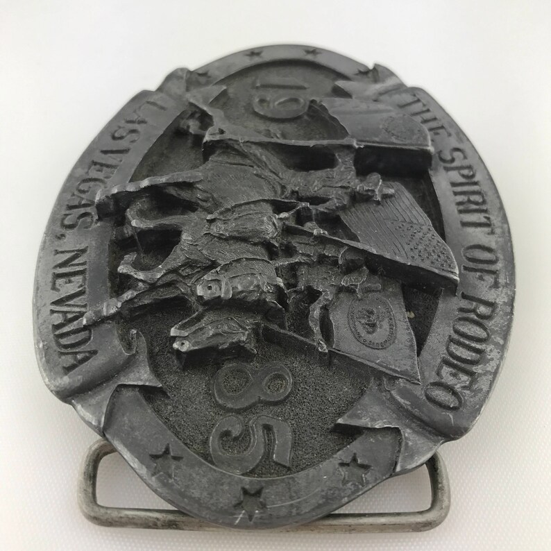 May include: A silver-colored metal belt buckle with a rodeo theme. The oval buckle features a detailed relief of a cowboy riding a bucking bronco, with the text "LAS VEGAS, NEVADA" and "THE SPIRIT OF RODEO". The number "85" is also visible.