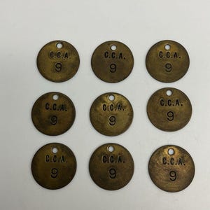 Könnte beinhalten: Neun runde Messinganhänger mit der Prägung "C.C.A." und der Zahl "9". Jeder Anhänger hat ein kleines Loch oben. Das Aussehen der Anhänger deutet darauf hin, dass sie alt sind.