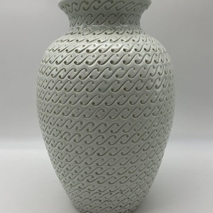 May include: A white ceramic vase with a rounded body and a flared rim. The exterior surface is covered in a repeating, embossed pattern of small, interconnected S-shaped curves and dots that create a textured, rhythmic visual effect.