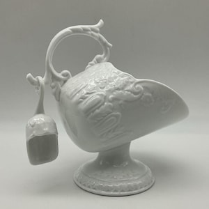 Op de afbeelding: Een witte porseleinen suikerpot met een handvat en een kleine lepel. De kom heeft een decoratief bloemmotief en staat op een voetstuk. De lepel hangt aan het handvat. Een klassiek stuk voor het serveren van suiker.