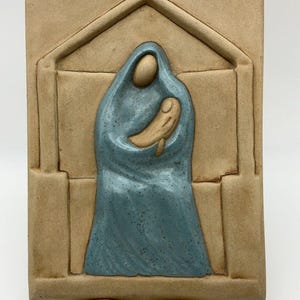 Puede incluir: Escultura en relieve de cerámica que representa una figura con una túnica azul sosteniendo otra más pequeña, enmarcada en un marco arquitectónico de color beige. La escultura está montada sobre un soporte de metal dorado. Las figuras son estilizadas.