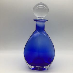 May include: A cobalt blue glass decanter with a clear glass stopper. The decanter has a rounded body and a long neck. The stopper is a clear, spherical shape. The decanter is likely used for storing and serving beverages.