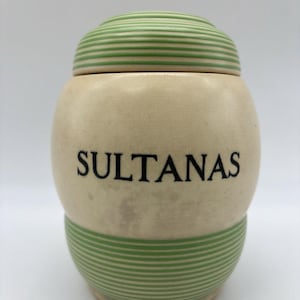 Puede incluir: Tarro de cerámica color crema con tapa, con rayas horizontales verdes en la tapa y la base. La palabra "SULTANAS" está impresa en negro en la parte delantera del tarro. El tarro tiene forma redondeada.