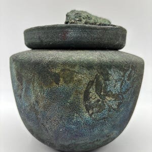 May include: A ceramic jar with a lid, featuring a mottled green and blue glaze with a crackled finish. The lid has a rough, textured surface.