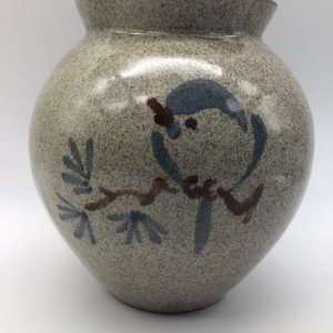May include: A speckled ceramic vase with a rounded body and a slightly flared rim. The vase features a hand-painted design of a blue bird perched on a brown branch with blue leaves. The overall color palette is neutral with brown and blue accents.