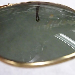 May include: A pair of sunglasses featuring dark, reflective lenses encased in a thin, polished gold-tone metal frame. The curved lenses show a clear reflection of the surrounding environment, including overhead lighting and a small dark loop detail.