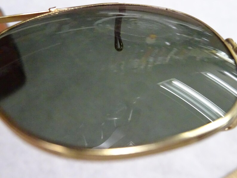 May include: A pair of sunglasses featuring dark, reflective lenses encased in a thin, polished gold-tone metal frame. The curved lenses show a clear reflection of the surrounding environment, including overhead lighting and a small dark loop detail.