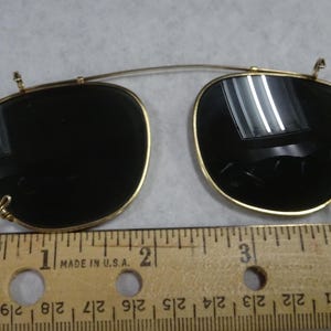 May include: A pair of gold-tone clip-on sunglasses with dark lenses and a thin metal bridge. The clip-on mechanism features small hooks on the sides and top. A wooden ruler is positioned underneath, showing the width is approximately 4.5 inches.