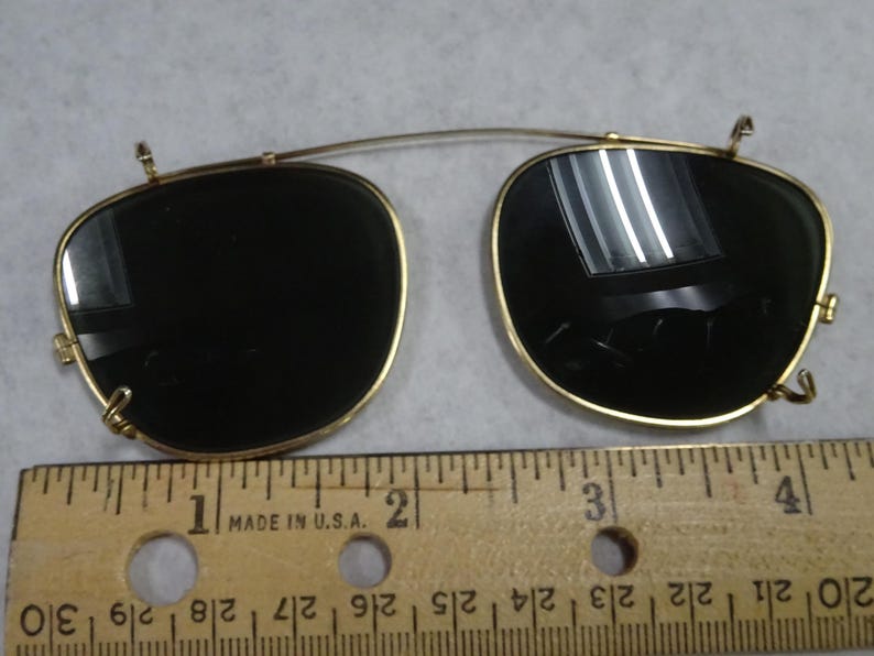May include: A pair of gold-tone clip-on sunglasses with dark lenses and a thin metal bridge. The clip-on mechanism features small hooks on the sides and top. A wooden ruler is positioned underneath, showing the width is approximately 4.5 inches.