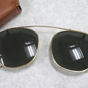 May include: A pair of gold-toned clip-on sunglasses with dark green lenses and a thin metal bridge connecting the two frames. The lenses are rounded and feature a reflective surface. A brown leather-textured case is partially visible in the corner.